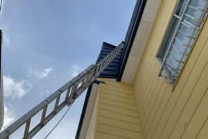 物置の屋根にはしごを立てかけ、高所の換気口へアプローチする様子。