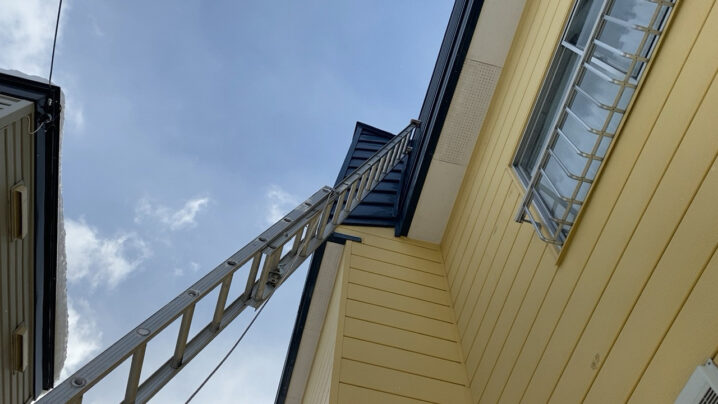 物置の屋根にはしごを立てかけ、高所の換気口へアプローチする様子。