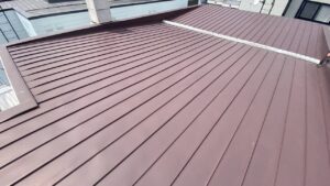 3階建ての建物の屋根の上に登り、平らなトタン板金の表面に剥がれや浮きが一切ないことを全体的に見渡して確認している点検中の写真