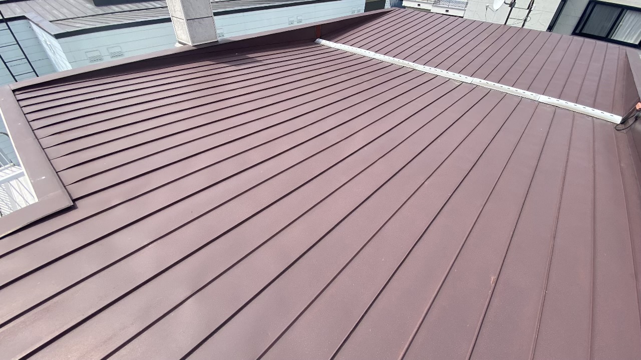 3階建ての建物の屋根の上に登り、平らなトタン板金の表面に剥がれや浮きが一切ないことを全体的に見渡して確認している点検中の写真