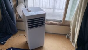 窓際に綺麗に設置された白いスポットクーラー本体と、窓パネルに繋がる蛇腹ホースの写真。リモコンが本体の上に置かれている