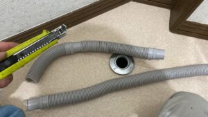 床の排水口の横で、余分に長かった洗濯機の排水ホースをカッターナイフを使って適切な長さに切断している様子