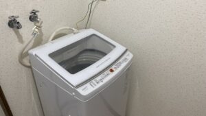 壁際にすっきりと設置され、電源が入りデジタル表示が灯っている状態のAQUA製の白い縦型洗濯機の全体写真