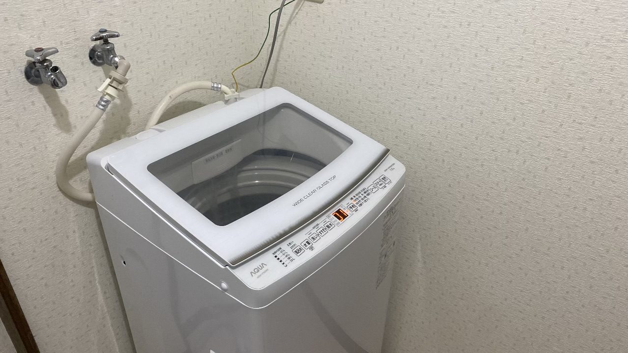 壁際にすっきりと設置され、電源が入りデジタル表示が灯っている状態のAQUA製の白い縦型洗濯機の全体写真