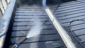 高圧洗浄機のノズルから勢いよく噴射される水煙とともに、黒い板金屋根の表面の目に見えない土埃やケレン粉塵を水洗いで吹き飛ばしている写真